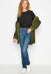 Czarna koszulka, oliwkowa kurtka z sznurkowego materiału, niebieskie dżinsy i białe trampki. Proste dżinsy z delikatnym przetarciem na dole.