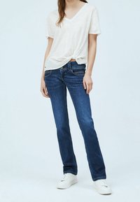 Kvinna klädd i vit knuten V-ringad t-shirt, mörkblå raka jeans och vita sneakers, stående mot en enkel bakgrund.