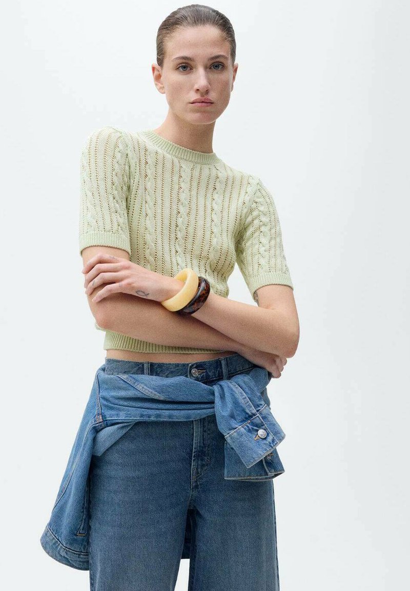 Un top tricotat cu mânecă scurtă, de culoare verde deschis, cu un model texturat, asortat cu blugi din denim cu talie înaltă și o jachetă din denim legată în jurul taliei.