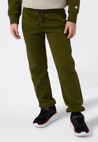 Ett barn som bär olivgröna sweatpants med dragsko och svarta sneakers med vita sulor och orangea detaljer, stående mot en vit bakgrund.