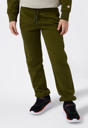 Barn som har på seg olivengrønne joggebukser med snøring og svarte sneakers med hvite såler og oransje detaljer, står mot en hvit bakgrunn.