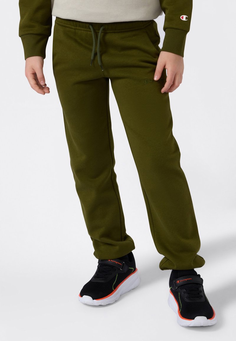 Ett barn som bär olivgröna sweatpants med dragsko och svarta sneakers med vita sulor och orangea detaljer, stående mot en vit bakgrund.