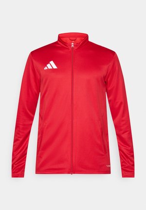 Rdeča jakna z zadrgo iz gladkega, lahkega blaga. Ima visok ovratnik in bel logotip na levi strani. Dva stranska žepa.