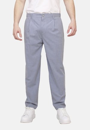 Uomo che indossa pantaloni azzurri su misura, sneakers bianchi e una camicia bianca, in piedi con le braccia rilassate lungo i fianchi.