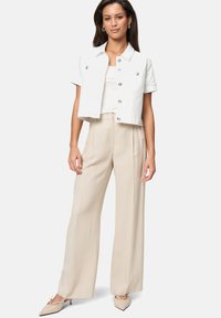 Vrouw in een witte, geknoopte, kortgestrikte jas met korte mouwen, beige wijde broek en beige puntige hakken, staand tegen een witte achtergrond.