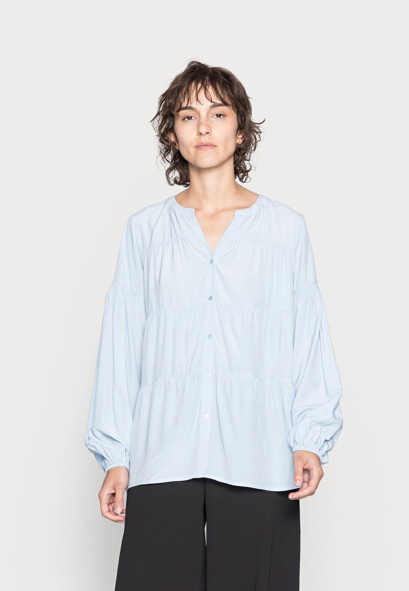Camisa azul clara, com decote redondo, camadas franzidas e mangas longas volumosas. Usada com calças pretas. Tecido com textura suave.