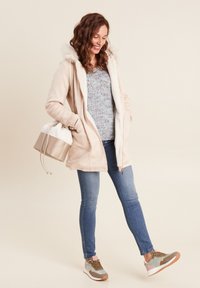 Lichtbeige jas met een zachte witte voering en capuchon, gecombineerd met een gestructureerde blauwe trui, blauwe skinny jeans en multikleurige sneakers.