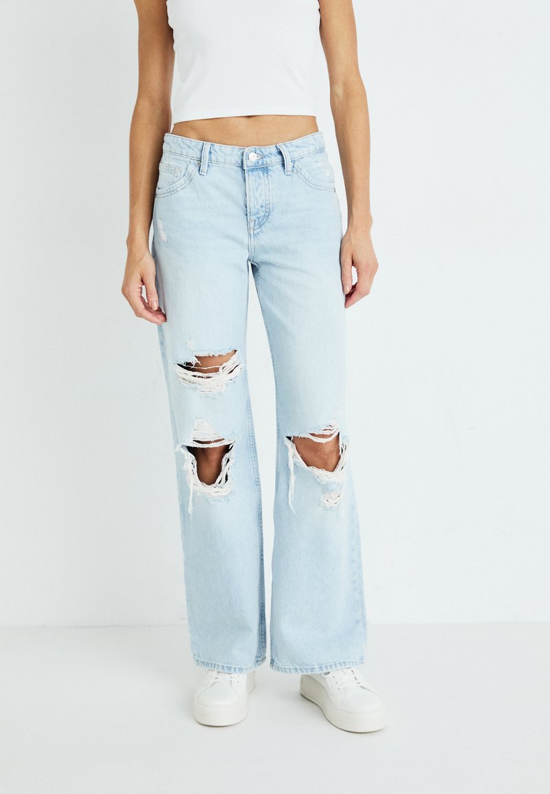 Svijetloplave široke traperice visokog struka s istaknutim rupama na koljenima, struganjem na rubovima i traper teksturom. Uparene s bijelim tenisicama.