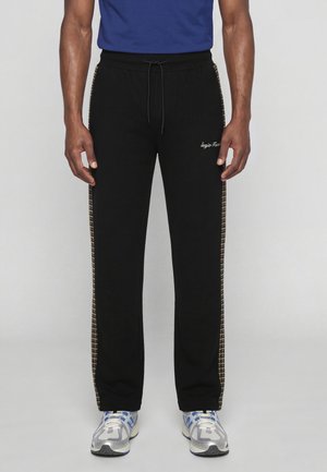 Homme portant un pantalon de survêtement noir avec des bandes latérales à carreaux, un logo blanc sur la cuisse, une chemise bleue et des chaussures de course blanches et bleues, debout.