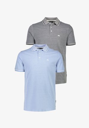 Uomo in piedi con polo azzurro, pantaloni neri e scarpe nere su sfondo bianco uniforme.