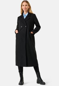 Cappotto lungo in misto lana nero con design doppiopetto, grandi bottoni e tasche laterali, abbinato a stivali neri con lacci e una camicia blu.