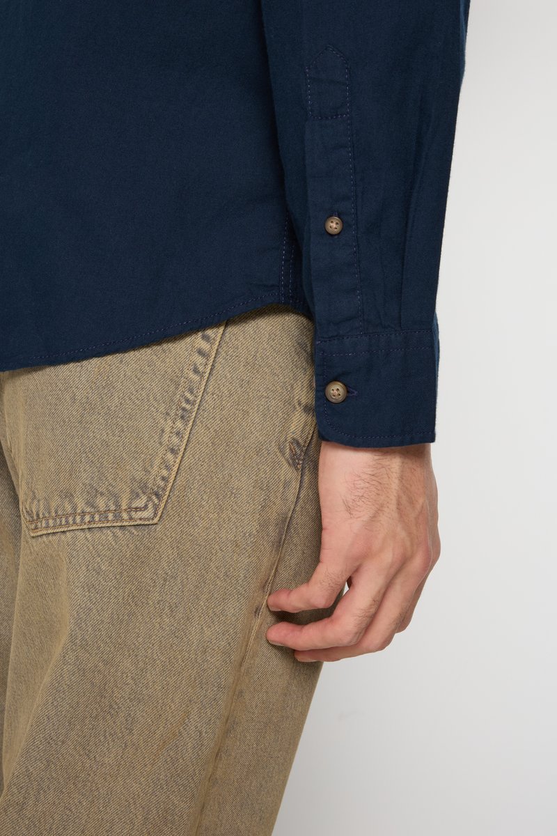 Camisa de botones de color azul marino con mangas largas, que presenta botones marrones, mostrada con pantalones beige texturizados. Detalle de costura visible en la tela.