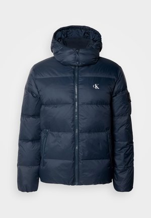 Marineblå pufferjakke med hette, glidelås og logoer. Har horisontale kviltepaneler og sidelommer. Laget av glatt, skinnende materiale.