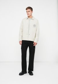 Giacca beige chiaro con bottoni a pressione e una tasca sul petto con logo a diamante, abbinata a pantaloni cargo neri e scarpe nere.