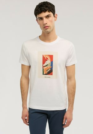 Joven con camiseta blanca que lleva una ilustración colorida de un perro y el texto "Trussardi", combinada con pantalones azul oscuro, posando sobre un fondo neutro.