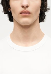 Κοντινό πλάνο από το κάτω μέρος προσώπου και τον λαιμό ενός ατόμου με κοντά σκούρα μαλλιά, που φοράει λευκό μπλουζάκι με στρογγυλή λαιμόκοψη και ένα μικρό κρίκο στο αριστερό αυτί.