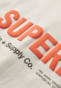 Tissu blanc arborant des lettres corail audacieuses portant le mot "SUPER" et un texte noir plus petit indiquant "Supply Co." L'impression texturée contraste avec le fond lisse.