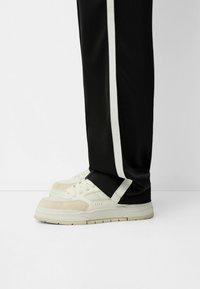 Světlé béžové tenisky s texturovaným textilním svrškem, bílé tkaničky a polstrovanou podrážkou, nošené s černými kalhotami s kontrastním bílým pruhem.