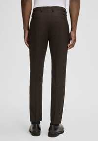 Braune maßgeschneiderte Hose mit glatter Textur, ausgestattet mit flacher Vorderseite, zwei Gesäßtaschen und einem konisch zulaufenden Bein. Getragen mit schwarzen Schuhen.