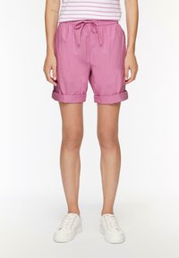 Shorts roses avec une taille élastique et un cordon de serrage, des revers roulés et un tissu léger. Portés avec des baskets blanches.