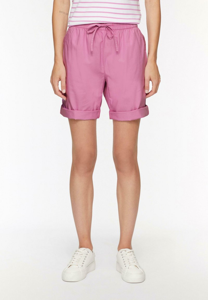 Shorts roses avec une taille élastique et un cordon de serrage, des revers roulés et un tissu léger. Portés avec des baskets blanches.