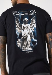 Schwarzes Baumwoll-T-Shirt mit einem grafischen Motiv einer geflügelten Statue vor einem blau-weißen Blitzhintergrund. "Closure Ldn"-Text über dem Bild.