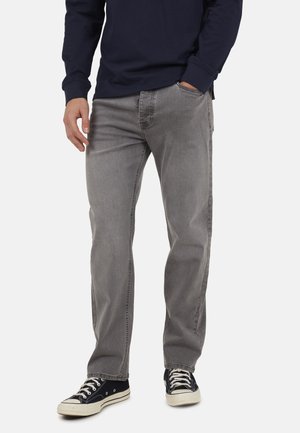 Man draagt grijze recht gesneden jeans, marineblauw longsleeve shirt en zwarte lage sneakers met witte veters, staat met één hand in zijn zak.