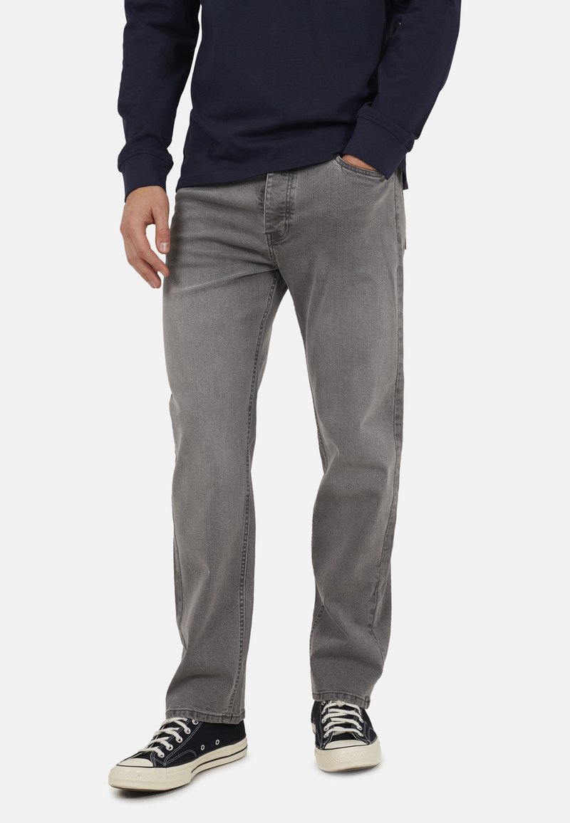 Mężczyzna ubrany w szare proste jeansy, granatową bluzę z długim rękawem i czarne niskie trampki z białymi sznurowadłami, stojący z jedną ręką w kieszeni.