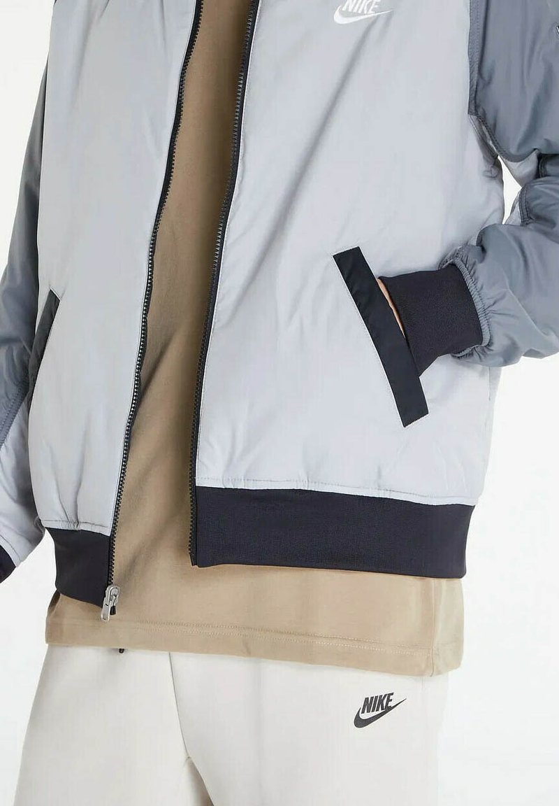 Primo piano di una persona che indossa una giacca Nike grigio chiaro, una camicia beige e pantaloni Nike bianchi, con una mano in tasca della giacca.