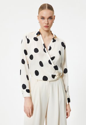 Blusa avvolgente crema con grandi pois neri, dotata di colletto e maniche lunghe, abbinata a pantaloni a vita alta.