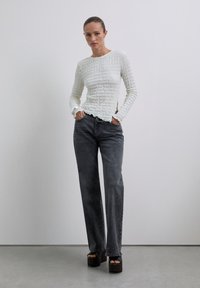 Kobieta w białej, strukturalnej bluzce z długim rękawem, w luźnych, czarnych jeansach o wysokim stanie oraz czarnych, platformowych sandałach na otwartym palcu, stoi z rękami w kieszeniach.