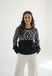 Czarnobiały sweter w wzory z frędzlami, zestawiony z jasnymi szerokimi spodniami. Biżuteria z turkusowymi akcentami.