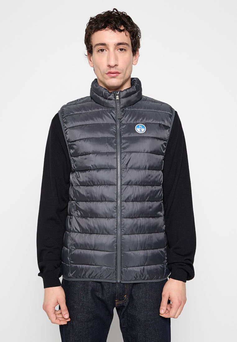 Gilet grigio imbottito con motivo a righe orizzontali e colletto alto, chiusura con zip sul davanti e una patch rotonda con logo sul lato sinistro. Sotto, una maglietta nera a maniche lunghe.