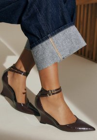 Bruine gepatineerde lederen slefhaks met een spitse neus en dubbele enkelbandjes. Jeans met een lichte denim omslag zijn gedeeltelijk zichtbaar.