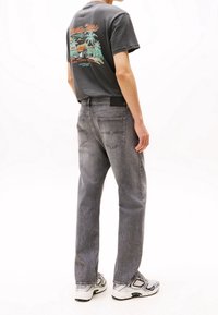 Young person wearing grey graphic t-shirt, faded grey straight-leg jeans, and white sneakers, standing with back and side visible.