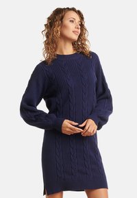 Abito maglione in blu navy con trama a treccia, maniche lunghe, orlo a coste e spacchi laterali. Realizzato in materiale morbido e caldo.
