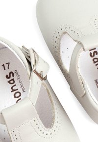 Zapatos Mary Jane de piel blanca que presentan detalles perforados, una hebilla ajustable de plata y un interior suave con una marca visible.