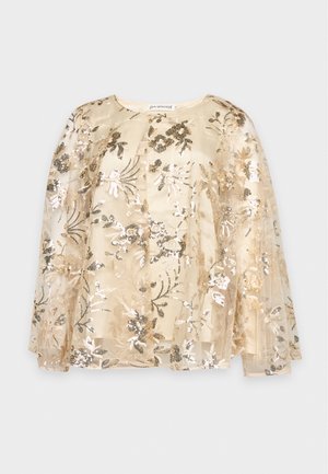 Le modèle se tient devant un fond de studio clair, vêtu d’un chemisier-cape transparent brodé de motifs floraux, d’un pantalon évasé taupe et de chaussures à talons pointues.