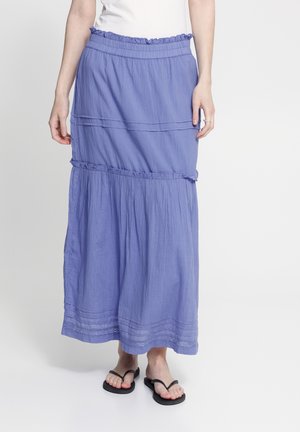 Donna che indossa una gonna lunga a balze color viola chiaro abbinata a infradito nere e top bianco, in piedi davanti a uno sfondo semplice.
