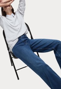 Camicia grigia a maniche lunghe con colletto e jeans in denim blu scuro loose-fit, seduto su una sedia nera contro uno sfondo bianco.