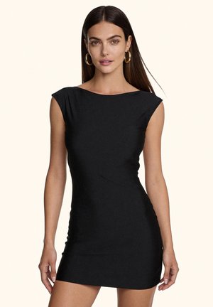 Giovane donna con lunghi capelli scuri che indossa un piccolo vestito nero senza maniche e orecchini a cerchio dorati, rivolta in avanti su uno sfondo uniforme.
