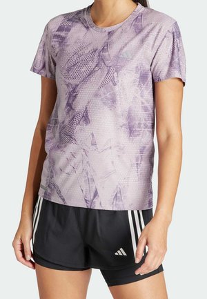 Vrouw draagt een paarse sportshirt met abstract patroon en korte mouwen en zwarte Adidas shorts met witte strepen en logo.