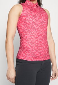 Vaaleanpunainen hihaton polo-paita, jossa on geometrinen valkoinen kuvio, kauluksellinen malli, slim fit -leikkaus ja kevyttä kangasta. Yhdistetty mustiin housuihin.