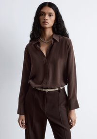 Brown button-up shirt in smooth fabric, featuring a collar, long sleeves, and a relaxed fit, paired with brown tailored pants and a light belt.