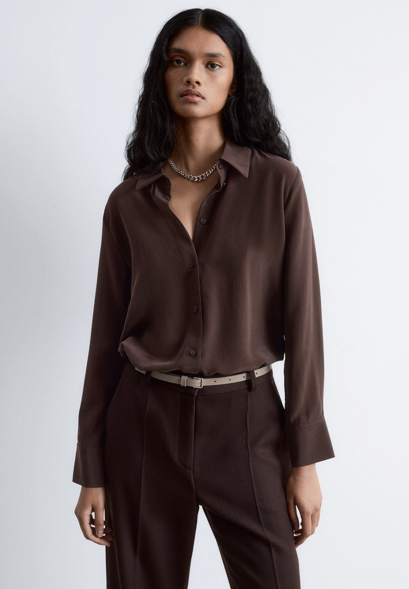 Brown button-up shirt in smooth fabric, featuring a collar, long sleeves, and a relaxed fit, paired with brown tailored pants and a light belt.