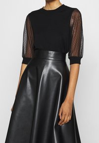 Svart topp med genomskinliga, prickiga meshärmar och rundad halslinning, kombinerad med en högmidjad svart fusk läderkjol, slät textur, A-linjeform.