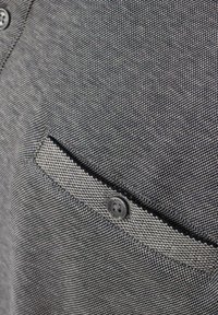 Grau Stoff mit strukturiertem Webmuster, ausgestattet mit einer kleinen Klappentasche und einem schwarzen Knopf mit Markenlogo. Das Design ist schlicht und funktional.