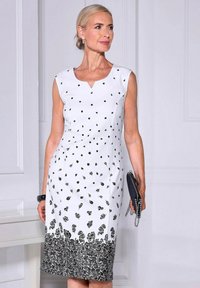 Weißes ärmelloses Kleid mit schwarzen Blumenmustern, V-Ausschnitt, strukturiertem Stoff und einem schwarzen Akzentsaum. Model hält eine schwarze Clutch.