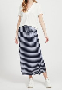 Maxi-jupe rayée bleu marine et blanc avec une fente latérale et une taille à cordon, associée à un chemisier blanc à manches courtes et des baskets blanches.