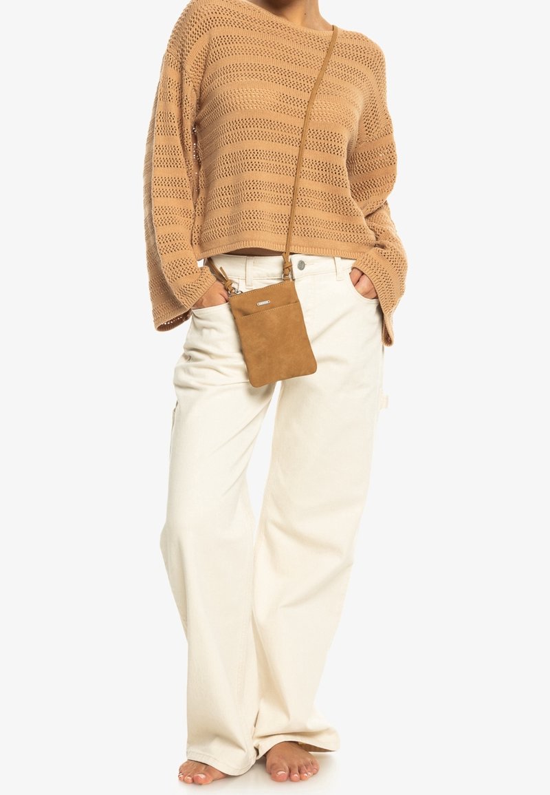 Beige stickad tröja med löst snitt och öppen stickning, kombinerad med vita vida byxor. En liten brun crossbody-väska hänger vid midjan.